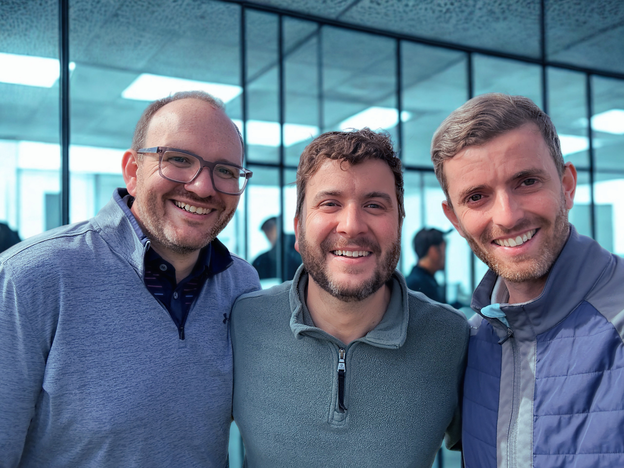 Three men smiling for a close-up group selfie in a glass-walled indoor space.